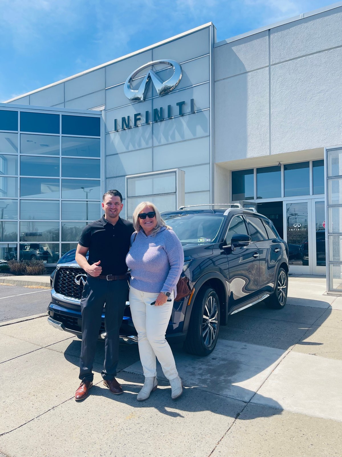 Two people standing beside a dark SUV in front of the INFINITI dealership building on a sunny day