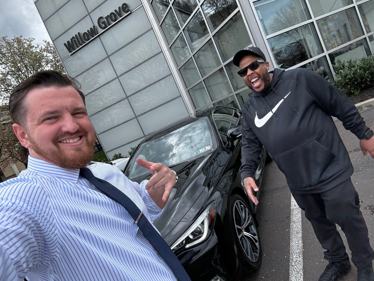 Two men posing with a black SUV in front of a building, both smiling at the camera