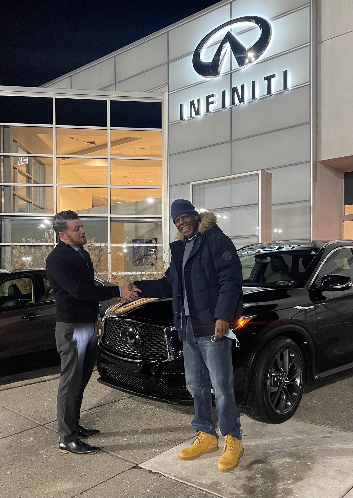 Two men shaking hands in front of a black luxury SUV at an Infiniti dealership showroom at night